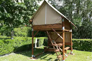 Zelt auf einer Holzplattform mit Leiter, im grünen Garten zwischen Bäumen und Hecken.