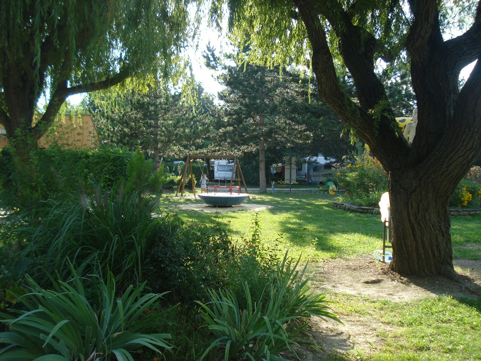 Campinggarten mit Rasenfläche, Wegen und großen Bäumen im Vordergrund, im Hintergrund Zelte und Wohnwagenstellplätze.