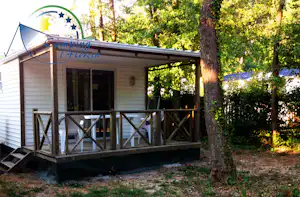 Camping-Lodge mit Veranda und Holzgeländer im Grünen, neben Bäumen und einem kleinen Weg am Stellplatz.
