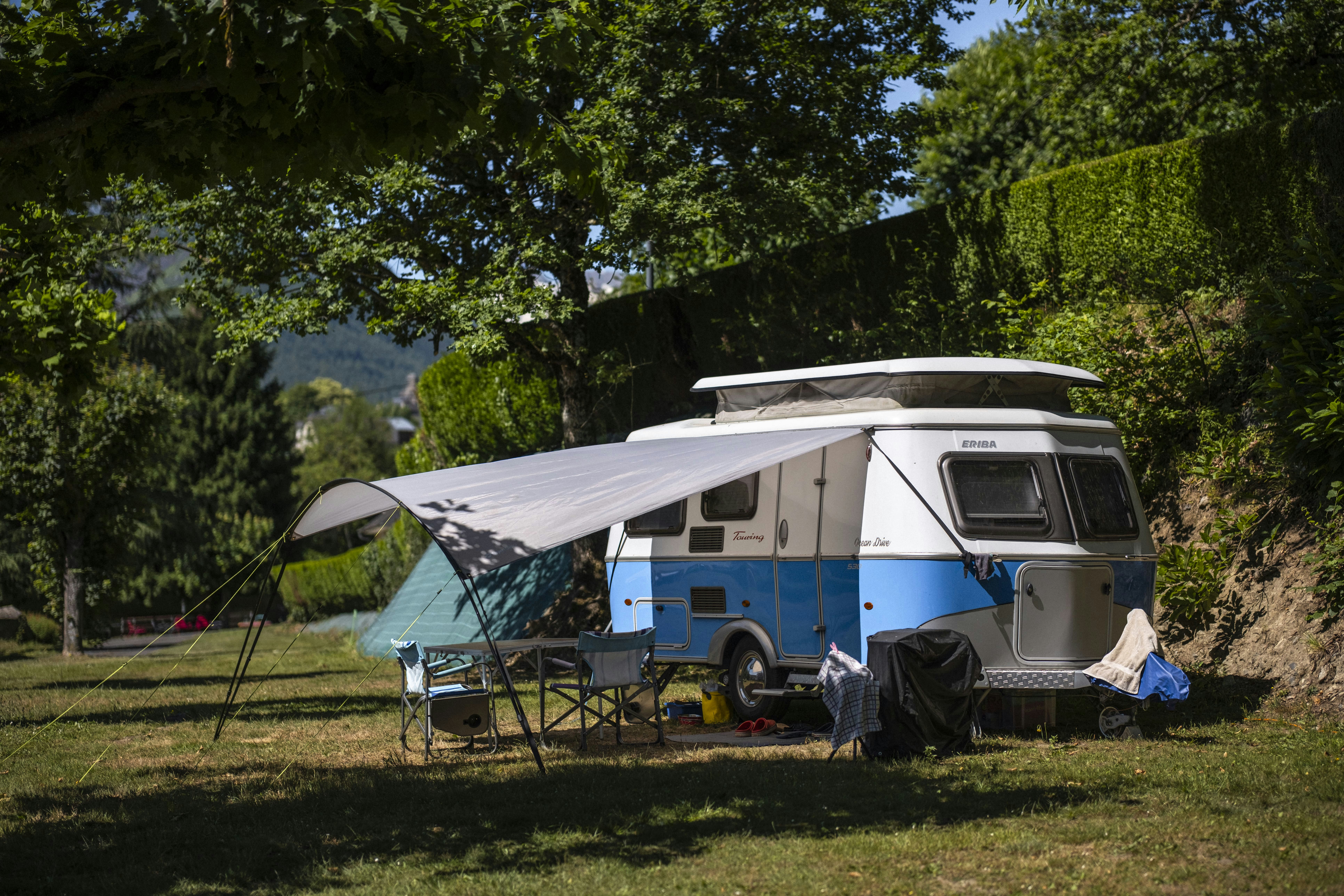 Wohnmobil mit Vorzelt und Campingstühlen auf einem Stellplatz im Grünen, umgeben von Bäumen und Blick in die Berge.