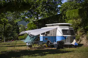 Wohnmobil mit Vorzelt und Campingstühlen auf einem Stellplatz im Grünen, umgeben von Bäumen und Blick in die Berge.