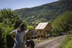 Eine Frau steht am Weg und blickt zu einem Holz-Unterstand mit Sonnensegel, umgeben von Bäumen und grünen Hügeln.