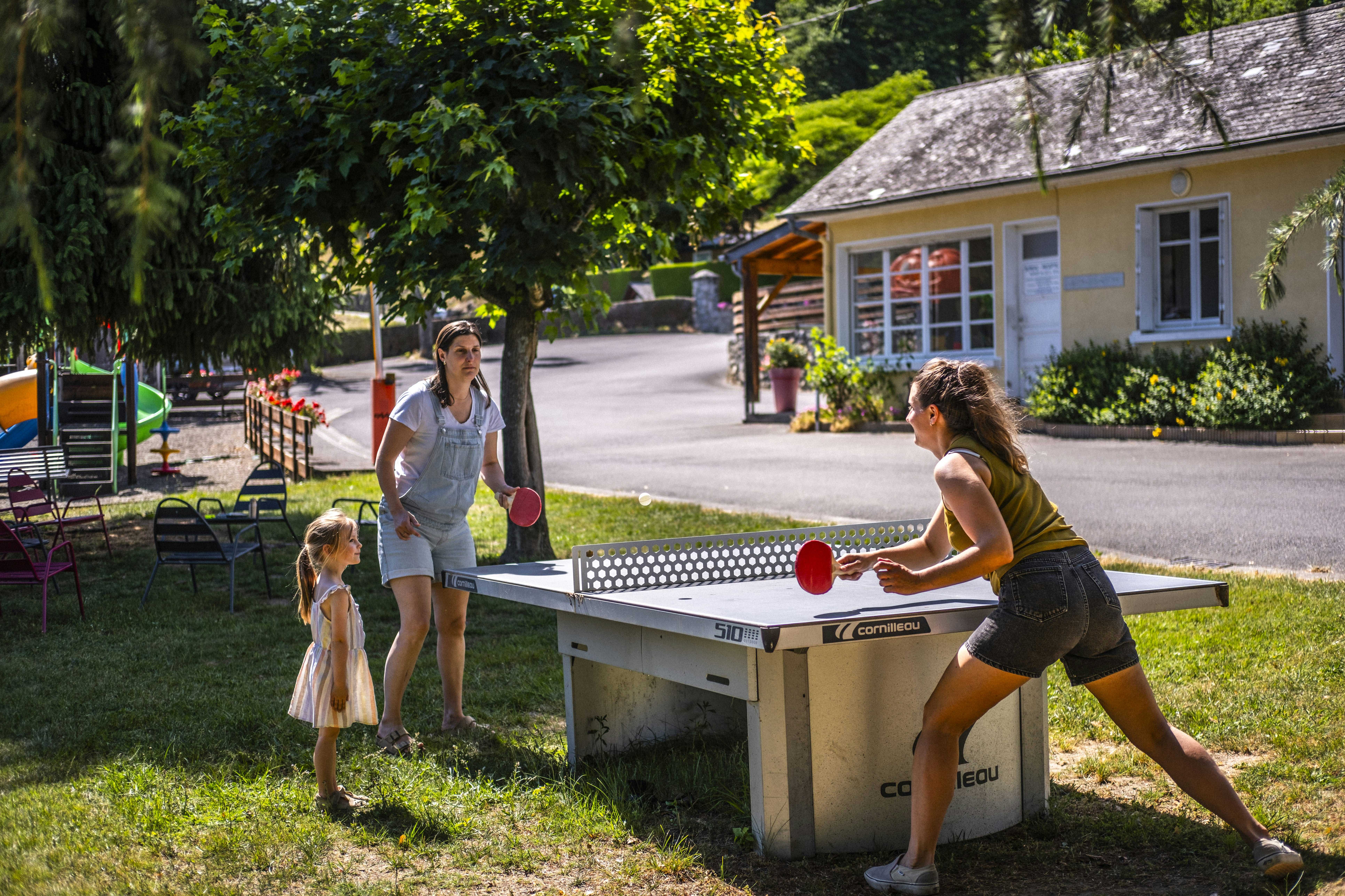 Familie spielt Tischtennis am Campingplatz vor einem gelben Gebäude, im Hintergrund Bäume und ein gepflasterter Weg.