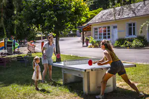 Familie spielt Tischtennis am Campingplatz vor einem gelben Gebäude, im Hintergrund Bäume und ein gepflasterter Weg.