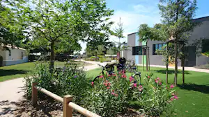 Blumenbeet mit Fahrrädern am Wegesrand vor einem modernen Gebäude, Rasen und Bäume auf dem Campingplatz.