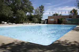 Schwimmbecken mit blauem Wasser, Betonrand und Liegen am Campingplatz, im Hintergrund Flaggen vor einem Gebäude.