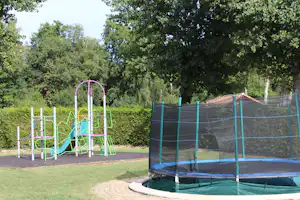Spielplatz mit buntem Klettergerüst und Trampolin auf einer Campingwiese, umgeben von Bäumen und Hecken.