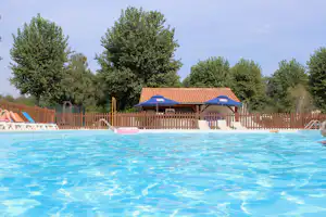 Campingpool mit blauem Wasser, Liegen und zwei Sonnenschirmen vor einem Holzpavillon, dahinter hohe Bäume und Zaun.