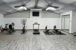 Fitnessraum mit mehreren Trainingsgeräten, Laufband und Fahrradergometer, großen Fenstern und weißem Hallendach.