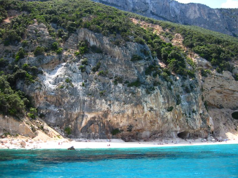 Türkisfarbenes Meer vor einer felsigen Küste mit bewaldeten Hängen und steilen Klippen im Hintergrund.