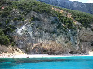 Türkisfarbenes Meer vor einer felsigen Küste mit bewaldeten Hängen und steilen Klippen im Hintergrund.