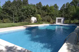 Schwimmbad mit blauem Wasser, Treppe am Beckenrand und Liegefläche; im Hintergrund Bäume, Rasen und ein weißes Gebäude.