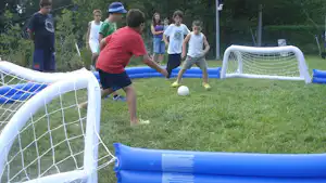 Kinder spielen Bubble-Soccer auf dem Campingplatz, mit blauen Toren und einem Ball auf dem Rasen.