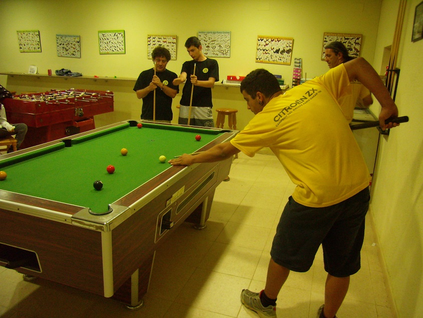 Billardtisch in einem Aufenthaltsraum, mehrere Spieler in gelben Shirts spielen, an der Wand bunte Poster und ein Tisch.