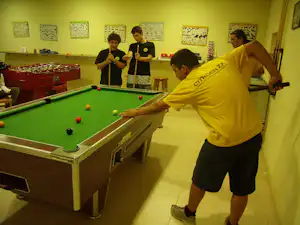 Billardtisch in einem Aufenthaltsraum, mehrere Spieler in gelben Shirts spielen, an der Wand bunte Poster und ein Tisch.