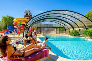 Familie am Pool auf Campingplatz, Liegen im Vordergrund, großes Glasdach über dem Schwimmbecken und Spielbereich im Hintergrund.