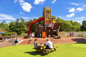 Spielplatz mit großer Rutsche und Kletterturm, daneben Rasenfläche und Picknicktisch mit zwei Personen.