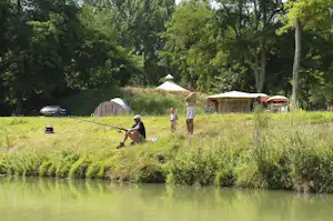 Zelte auf einer Wiese am Fluss, im Vordergrund ein Kind am Ufer und weitere Personen zwischen Bäumen.