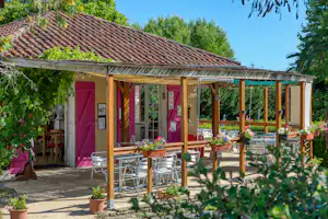 Terrasse mit Holzdach und Sitzgruppe vor einem Ferienhaus mit roten Türen und Fenstern, umgeben von Bäumen und Pflanzen.