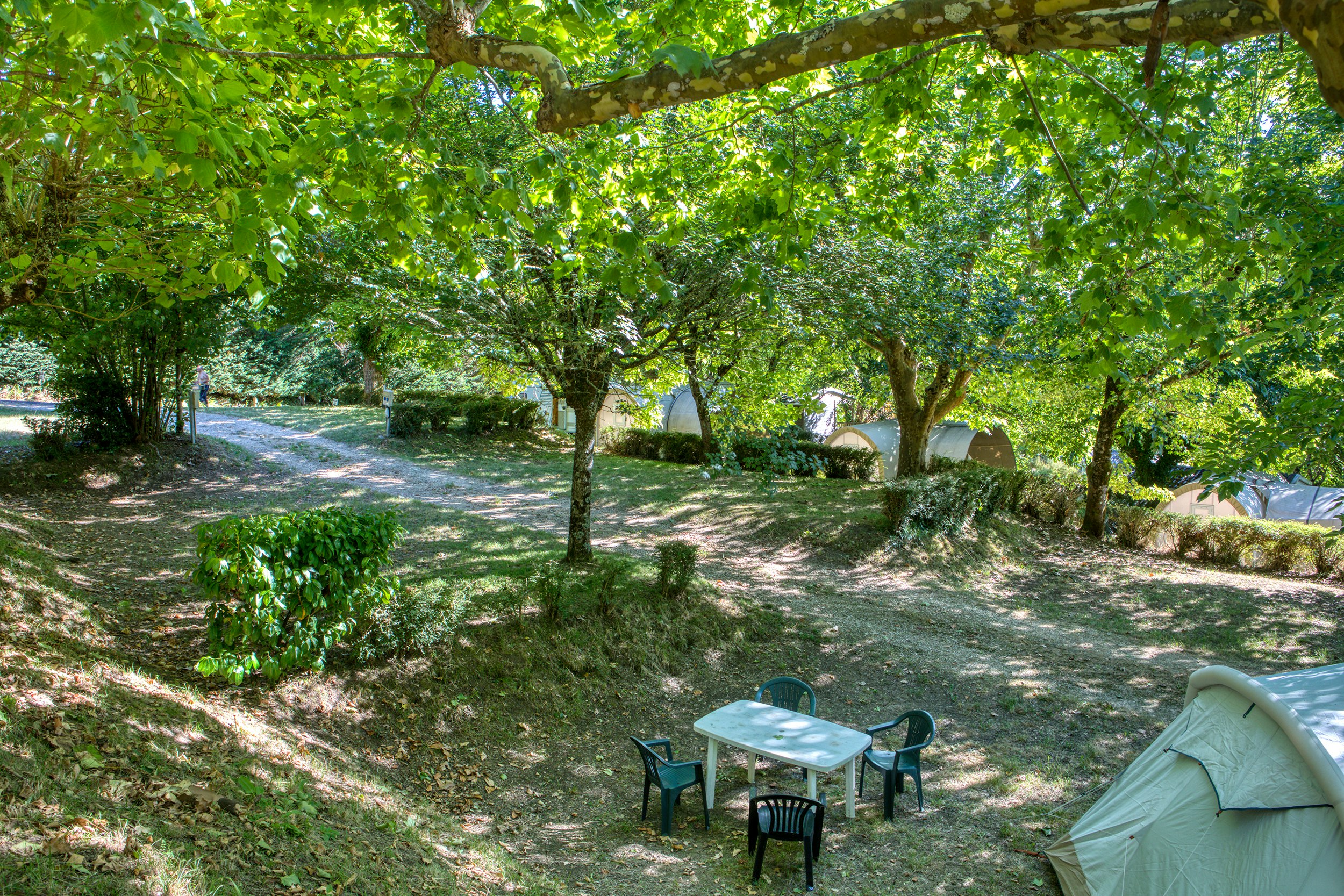 Grüner Campingplatz im Schatten von Bäumen, mit Zelt und kleinen Gartenmöbeln auf einem Kiesweg.