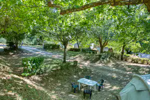Grüner Campingplatz im Schatten von Bäumen, mit Zelt und kleinen Gartenmöbeln auf einem Kiesweg.