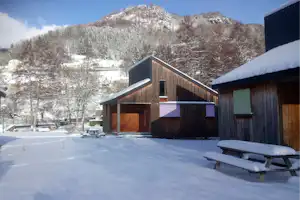 Holz-Hütte mit verschneitem Dach, Bank und Sitzbereich vor dem Gebäude, im Hintergrund schneebedeckte Berge und Bäume.