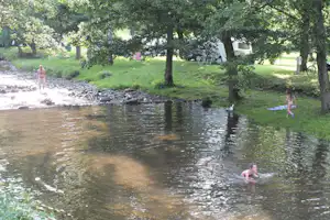 Bach im grünen Wald mit Personen im Wasser, Zelte und Campingstühle am Ufer, Bäume und Rasen im Hintergrund.
