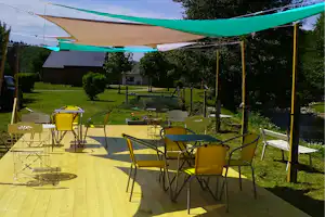 Gelbe Campingstühle und Tisch auf der Terrasse, Sonnensegel über dem Platz, grüner Garten mit Bäumen und Gartenhaus im Hintergrund.