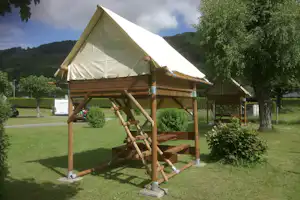Zelt mit Holzplattform und Leiter auf einem Campingplatz, umgeben von Rasen, Bäumen und Hügeln im Hintergrund.