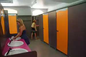 Waschraum im Campingbereich mit mehreren Waschbecken, orangefarbenen Kabinentüren und Fliesenboden.