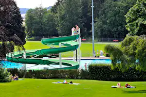 Grüne Wasserrutschen am Pool, daneben Liegewiese mit Bäumen und Zaun im Campinggelände.