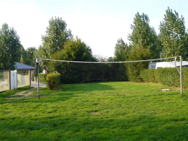 Grüner Rasen auf einem Campingplatz mit Zaun, Hecke und Bäumen im Hintergrund, daneben ein Gebäude und ein Weg.