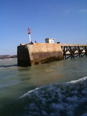 Wellen vor einer steinernen Seemauer mit Leuchtturm, dahinter ein kleines Gebäude und ein Holzsteg.