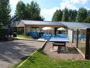 Terrasse am Campingplatz mit Gartenmöbeln, mehreren blauen Liegestühlen und Tisch vor einem Holzgebäude, umgeben von Bäumen.