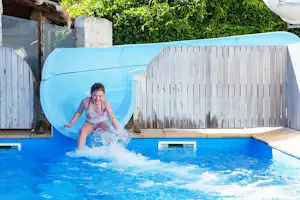 Kind planscht im blauen Pool vor einer weißen Holzumrandung und einem aufblasbaren Wasserrutscheneinsatz im Garten.