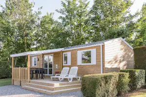 Holzbungalow mit Terrasse, weißen Fensterläden und zwei Liegestühlen vor Hecken und Bäumen im Campingbereich.