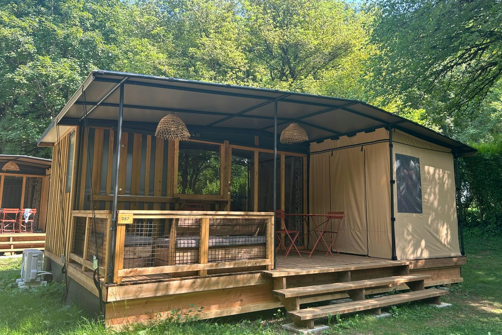 Campingunterkunft mit Veranda und Holzpodest, seitlich Vorhänge und Sitzbank, im Grünen zwischen Bäumen.