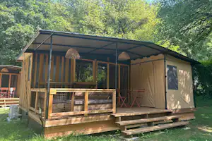 Campingunterkunft mit Veranda und Holzpodest, seitlich Vorhänge und Sitzbank, im Grünen zwischen Bäumen.