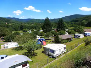 Campingwiese mit Wohnwagen und Zelten, Bäume und Hecken, im Hintergrund Berge und ein See.