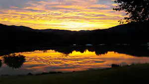 Sonnenuntergang über einem See im Tal, mit Spiegelung am Wasser, Hügeln und dunklen Bäumen am Ufer.