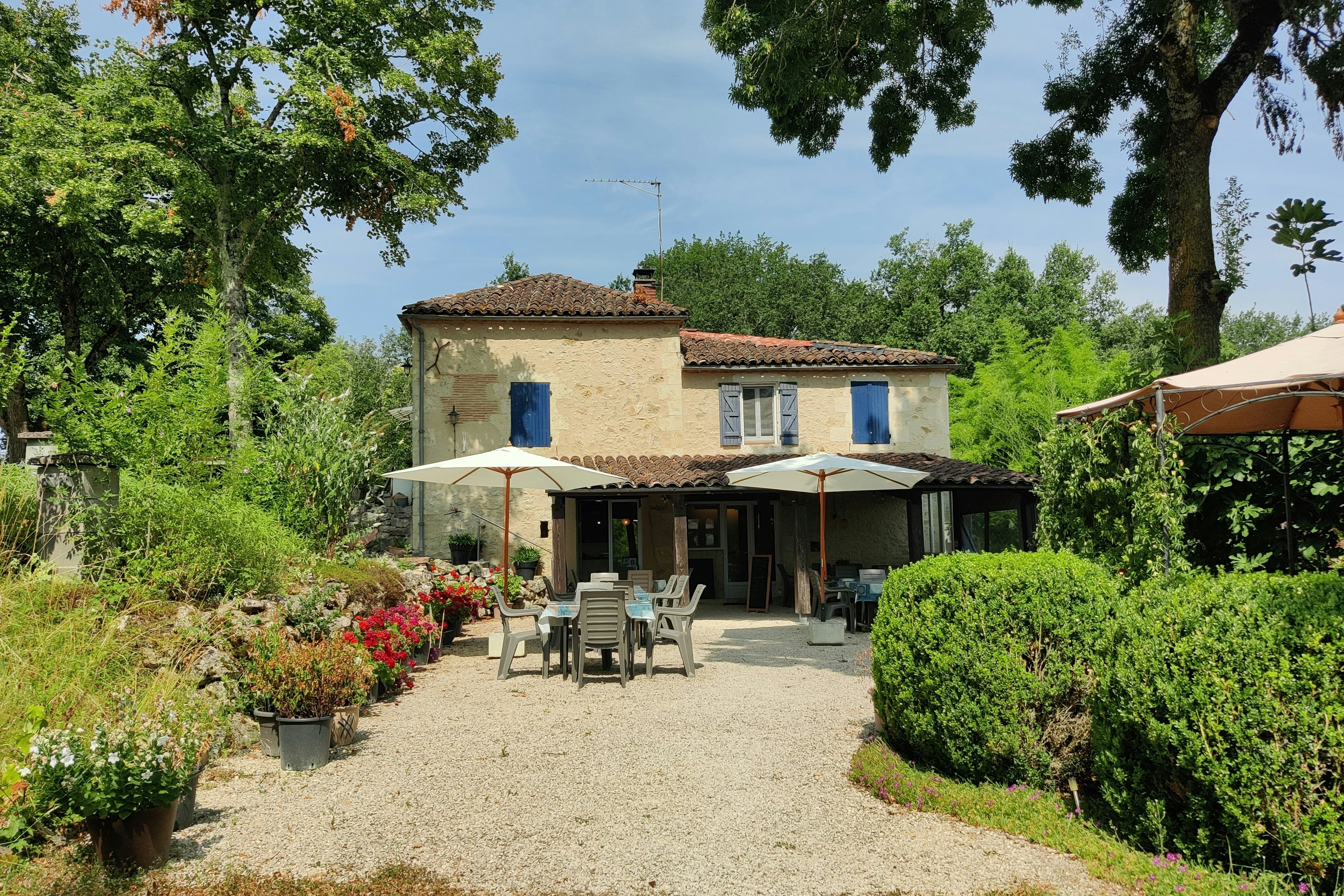 Campinghaus mit Terrasse, Sonnenschirmen und Gartenmöbeln vor gepflegten Hecken und Blumenbeeten, umgeben von Bäumen.