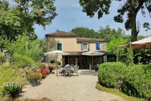 Campinghaus mit Terrasse, Sonnenschirmen und Gartenmöbeln vor gepflegten Hecken und Blumenbeeten, umgeben von Bäumen.