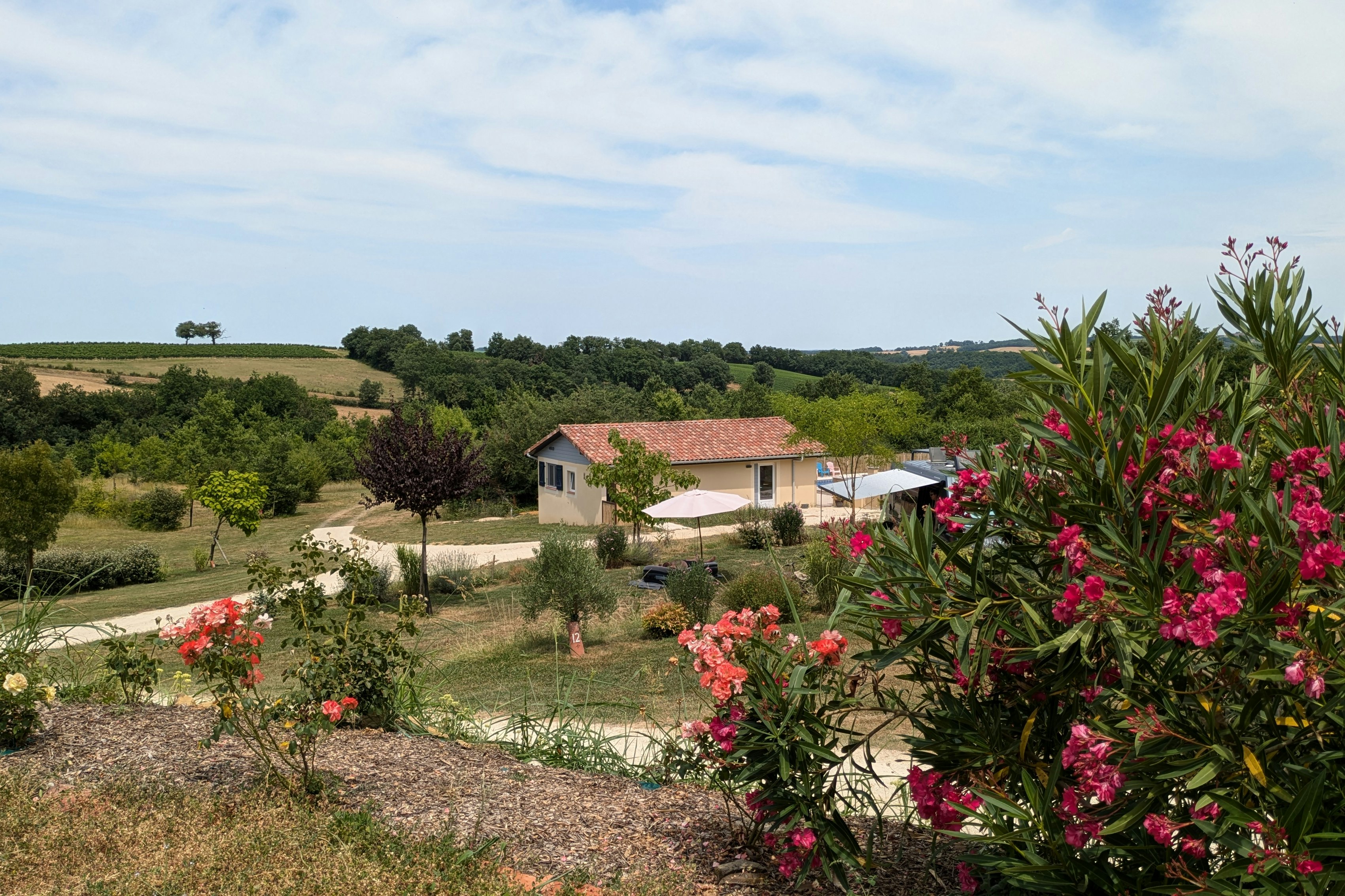 Campinghaus mit rotem Dach, umgeben von Wegen und blühenden Sträuchern, im Hintergrund grüne Hügel und Bäume.