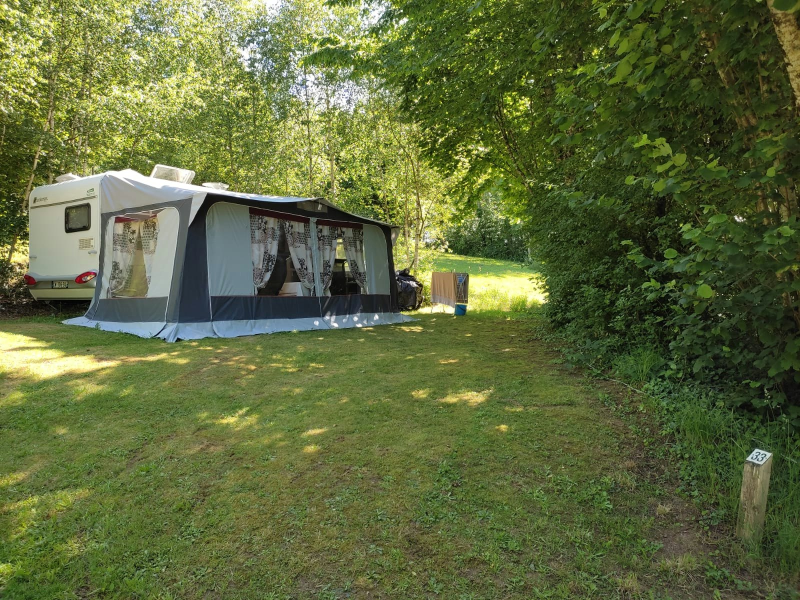 Campingzelt mit Vorzelt und Wohnwagen-Anbau auf einem Grasstellplatz, umgeben von Bäumen und Hecken im Grünen.