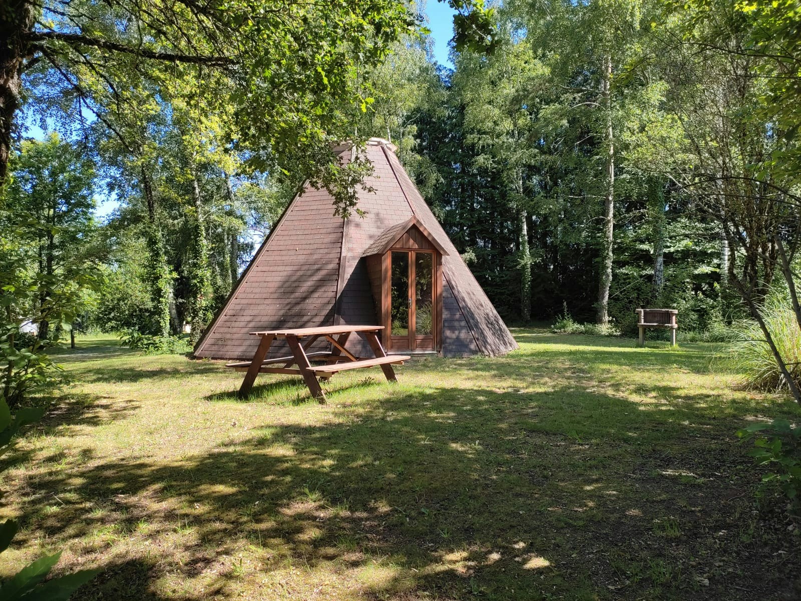Campingzelt mit Holzveranda im Grünen, umgeben von Bäumen und Rasenfläche auf einem Campingplatz.