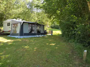 Wohnwagen mit Vorzelt auf einer Campingwiese, daneben ein grüner Müllbehälter und Bäume im Hintergrund.