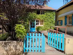 Haus am Campingplatz mit blauem Holzzaun, Terrasse und grüner Hecke vor den Fenstern, umgeben von Bäumen.