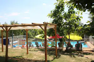 Poolbereich am Campingplatz mit Pergola, Liegestühlen und Sonnenschirmen, umgeben von Rasen und Bäumen.