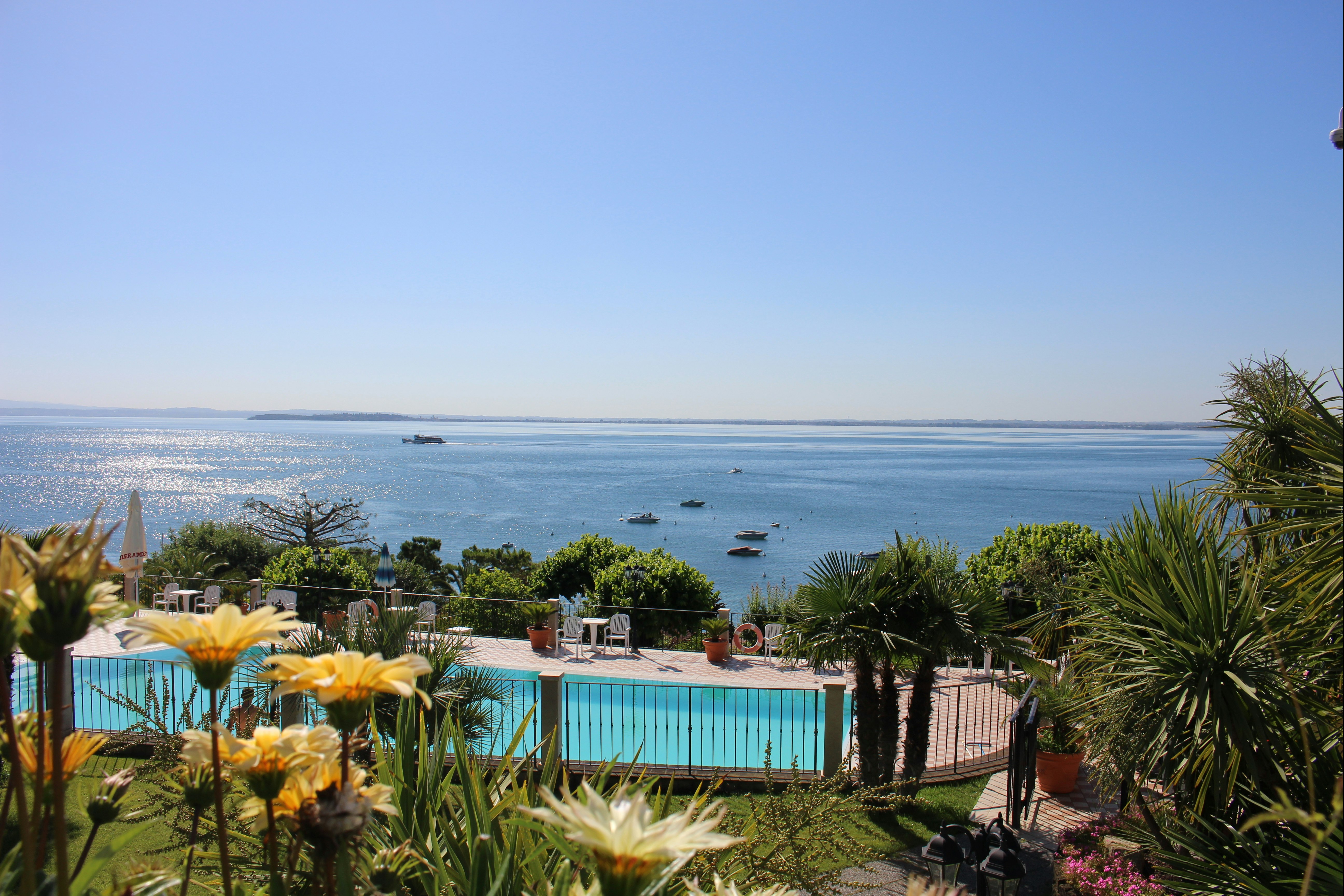 Blick vom Campingplatz auf das Meer mit blauem Pool, Palmen und Blumenbeeten im Vordergrund.