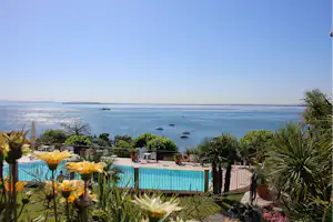Blick vom Campingplatz auf das Meer mit blauem Pool, Palmen und Blumenbeeten im Vordergrund.